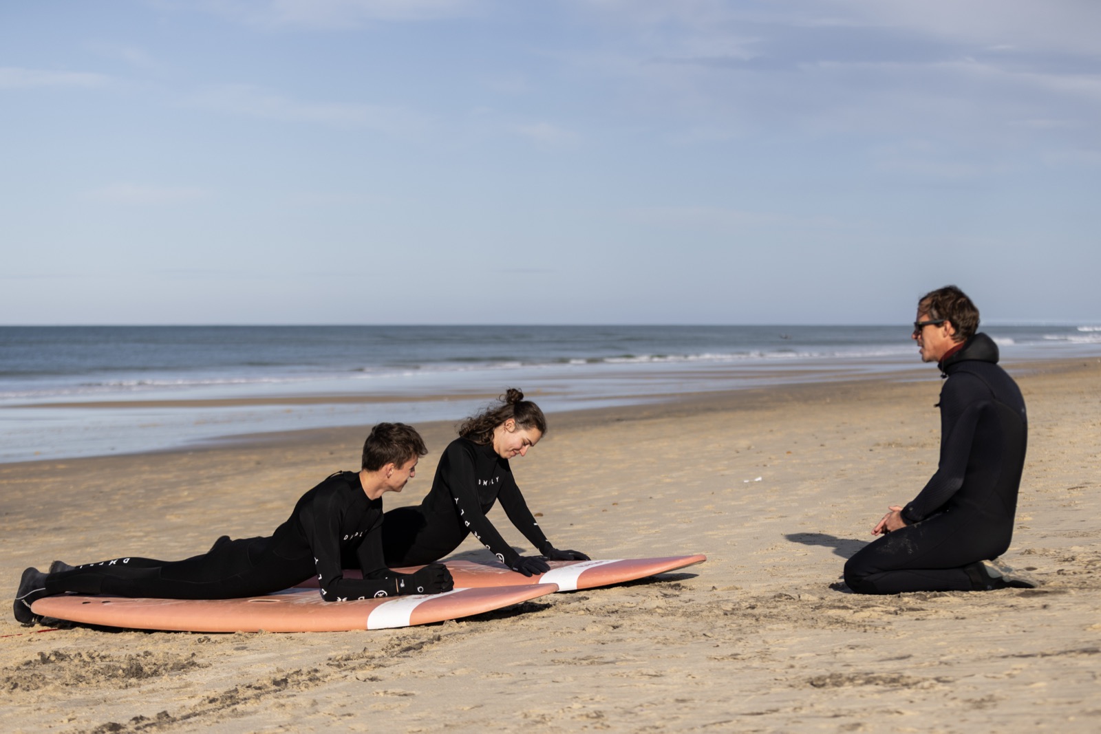 Sur le sable, deux élèves allongés sur leur planche écoutent les conseils de Sylvain